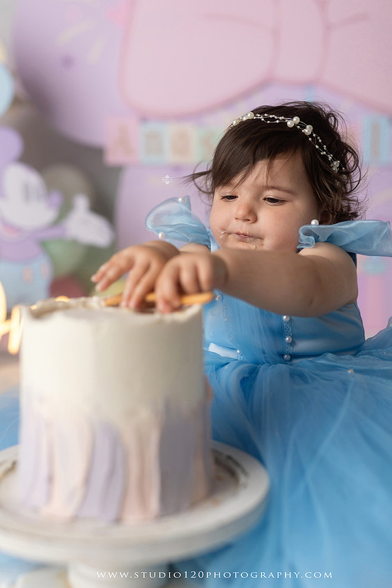 Disneyland-inspired first birthday cake smash featuring a baby girl in a blue princess dress with pastel balloons, whimsical amusement park décor, custom birthday backdrop, milestone portraits, and cake smash photography by Studio 120 Photography in Ramona, serving San Diego County.