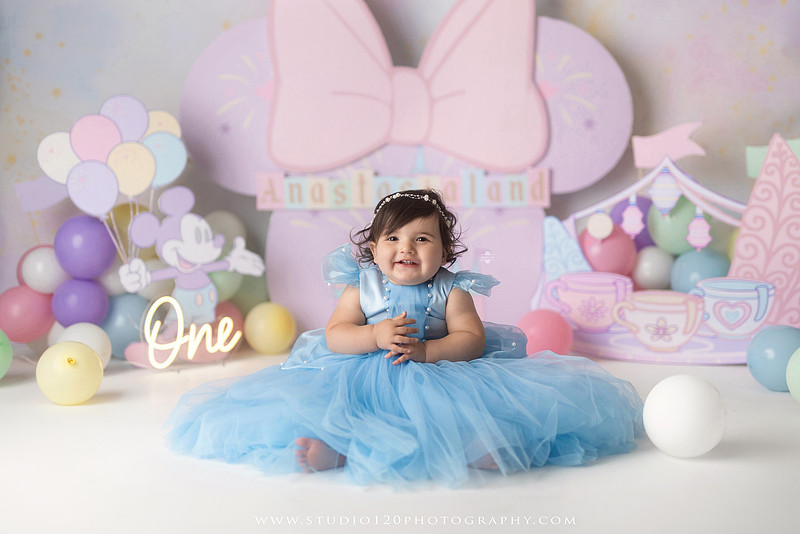 Disneyland-inspired first birthday cake smash featuring a baby girl in a blue princess dress with pastel balloons, whimsical amusement park décor, custom birthday backdrop, milestone portraits, and cake smash photography by Studio 120 Photography in Ramona, serving San Diego County.