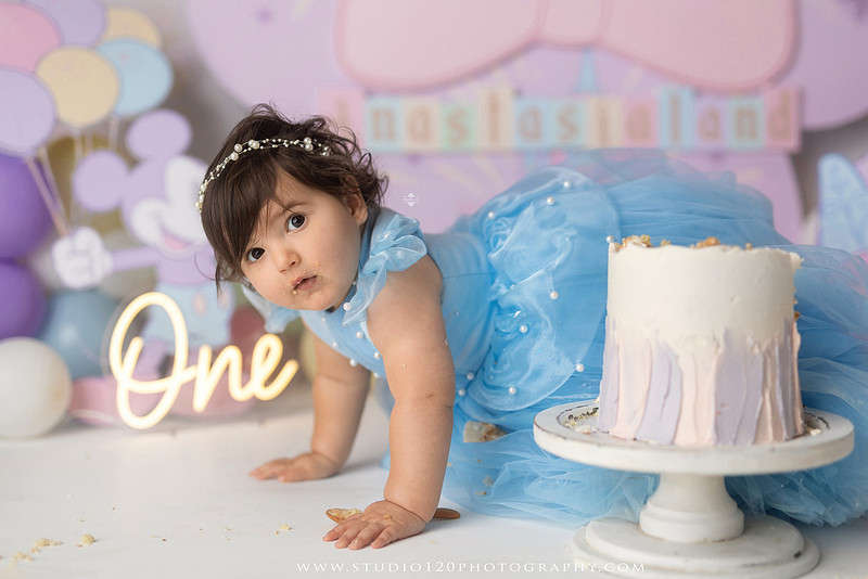Disneyland-inspired first birthday cake smash featuring a baby girl in a blue princess dress with pastel balloons, whimsical amusement park décor, custom birthday backdrop, milestone portraits, and cake smash photography by Studio 120 Photography in Ramona, serving San Diego County.