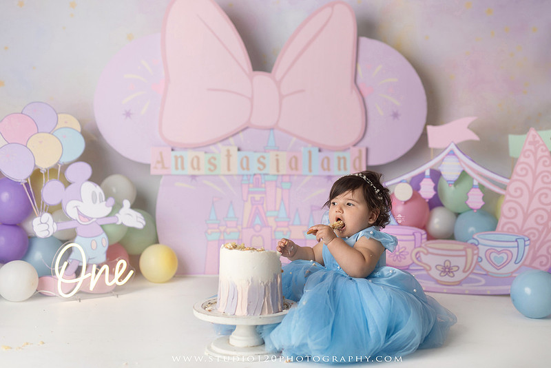 Disneyland-inspired first birthday cake smash featuring a baby girl in a blue princess dress with pastel balloons, whimsical amusement park décor, custom birthday backdrop, milestone portraits, and cake smash photography by Studio 120 Photography in Ramona, serving San Diego County.