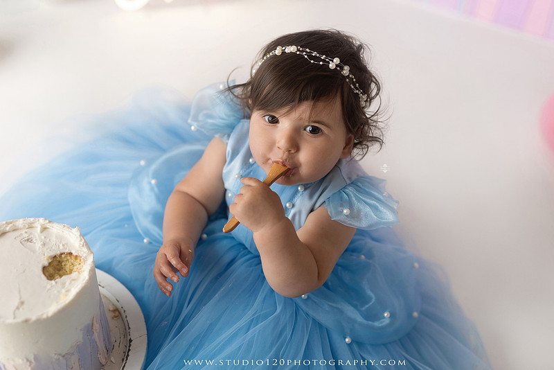 Disneyland-inspired first birthday cake smash featuring a baby girl in a blue princess dress with pastel balloons, whimsical amusement park décor, custom birthday backdrop, milestone portraits, and cake smash photography by Studio 120 Photography in Ramona, serving San Diego County.