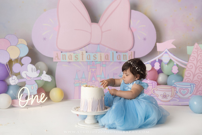 Disneyland-inspired first birthday cake smash featuring a baby girl in a blue princess dress with pastel balloons, whimsical amusement park décor, custom birthday backdrop, milestone portraits, and cake smash photography by Studio 120 Photography in Ramona, serving San Diego County.