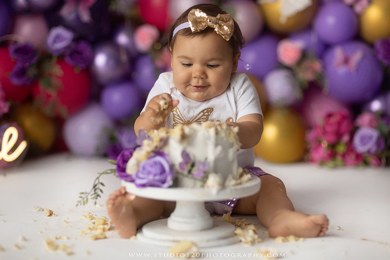 Colorful butterfly-themed first birthday cake smash featuring a happy baby girl enjoying birthday cake surrounded by vibrant purple, pink, and gold balloons, floral accents, butterflies, and a glowing "One" sign at Studio 120 Photography in Ramona, California.