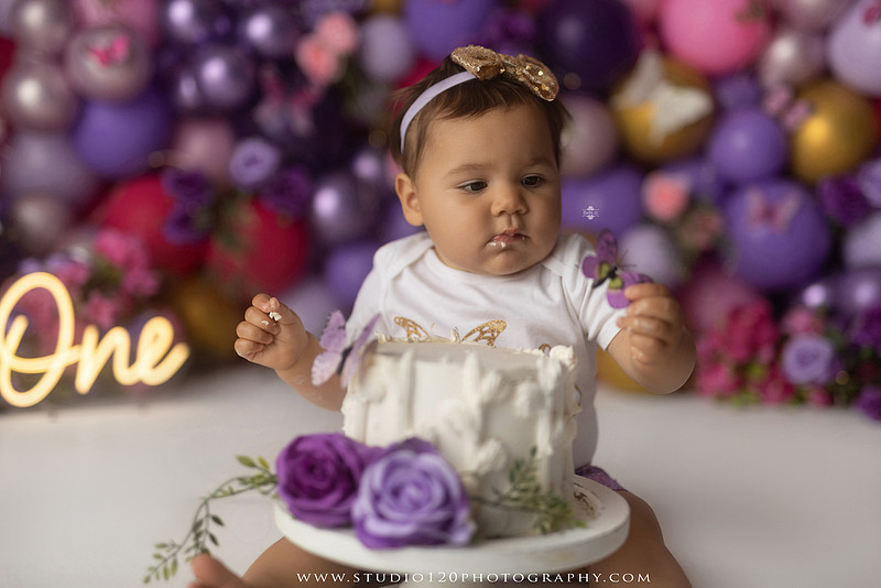 Colorful butterfly-themed first birthday cake smash featuring a happy baby girl enjoying birthday cake surrounded by vibrant purple, pink, and gold balloons, floral accents, butterflies, and a glowing "One" sign at Studio 120 Photography in Ramona, California.
