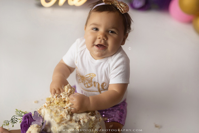 Colorful butterfly-themed first birthday cake smash featuring a happy baby girl enjoying birthday cake surrounded by vibrant purple, pink, and gold balloons, floral accents, butterflies, and a glowing "One" sign at Studio 120 Photography in Ramona, California.