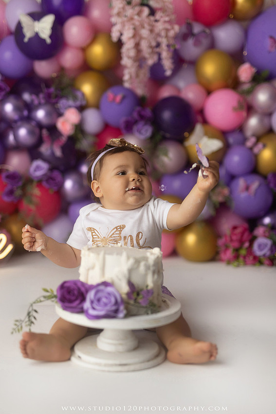Colorful butterfly-themed first birthday cake smash featuring a happy baby girl enjoying birthday cake surrounded by vibrant purple, pink, and gold balloons, floral accents, butterflies, and a glowing "One" sign at Studio 120 Photography in Ramona, California.
