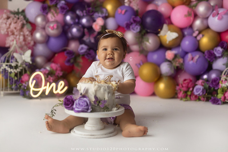 Colorful butterfly-themed first birthday cake smash featuring a happy baby girl enjoying birthday cake surrounded by vibrant purple, pink, and gold balloons, floral accents, butterflies, and a glowing "One" sign at Studio 120 Photography in Ramona, California.