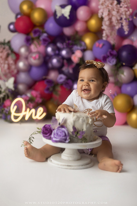 Colorful butterfly-themed first birthday cake smash featuring a happy baby girl enjoying birthday cake surrounded by vibrant purple, pink, and gold balloons, floral accents, butterflies, and a glowing "One" sign at Studio 120 Photography in Ramona, California.
