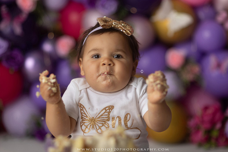 Colorful butterfly-themed first birthday cake smash featuring a happy baby girl enjoying birthday cake surrounded by vibrant purple, pink, and gold balloons, floral accents, butterflies, and a glowing "One" sign at Studio 120 Photography in Ramona, California.