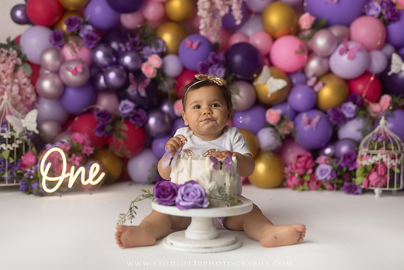 Colorful butterfly-themed first birthday cake smash featuring a happy baby girl enjoying birthday cake surrounded by vibrant purple, pink, and gold balloons, floral accents, butterflies, and a glowing "One" sign at Studio 120 Photography in Ramona, California.