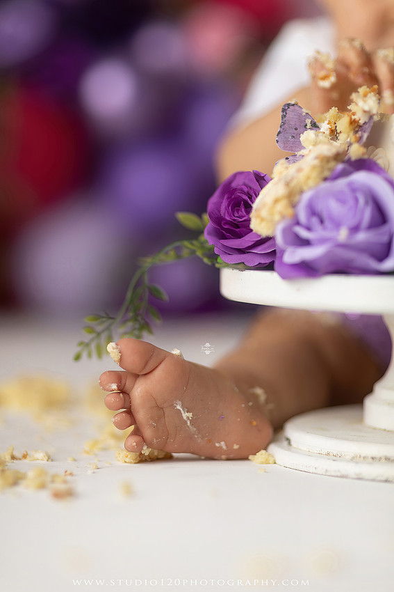 Colorful butterfly-themed first birthday cake smash featuring a happy baby girl enjoying birthday cake surrounded by vibrant purple, pink, and gold balloons, floral accents, butterflies, and a glowing "One" sign at Studio 120 Photography in Ramona, California.