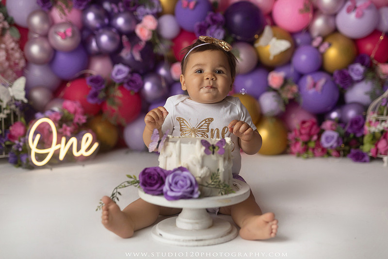 Colorful butterfly-themed first birthday cake smash featuring a happy baby girl enjoying birthday cake surrounded by vibrant purple, pink, and gold balloons, floral accents, butterflies, and a glowing "One" sign at Studio 120 Photography in Ramona, California.