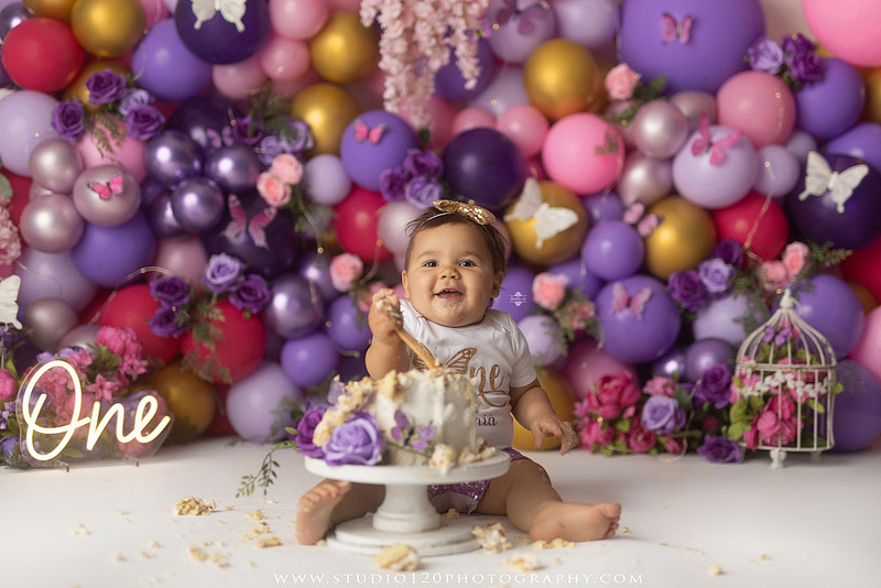 Colorful butterfly-themed first birthday cake smash featuring a happy baby girl enjoying birthday cake surrounded by vibrant purple, pink, and gold balloons, floral accents, butterflies, and a glowing "One" sign at Studio 120 Photography in Ramona, California.