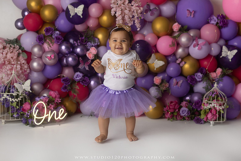 Colorful butterfly-themed first birthday cake smash featuring a happy baby girl enjoying birthday cake surrounded by vibrant purple, pink, and gold balloons, floral accents, butterflies, and a glowing "One" sign at Studio 120 Photography in Ramona, California.