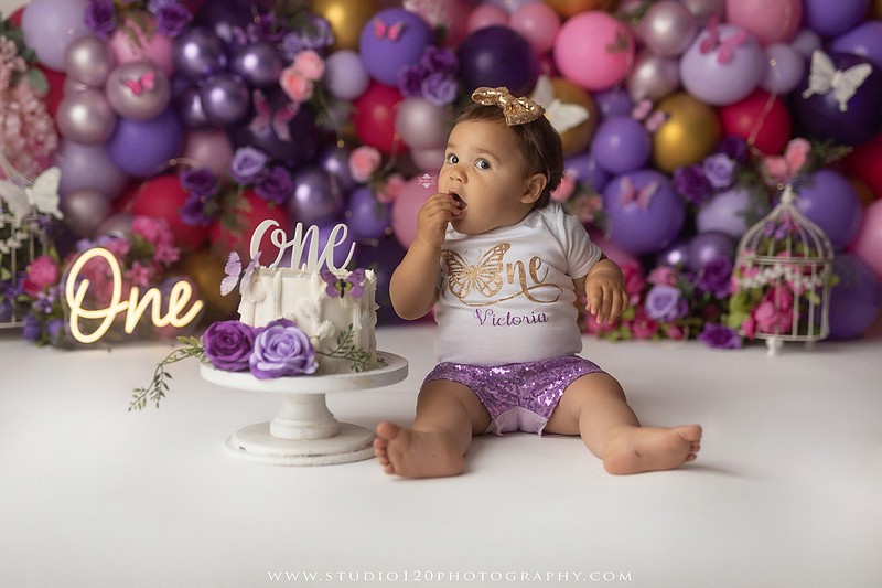 Colorful butterfly-themed first birthday cake smash featuring a happy baby girl enjoying birthday cake surrounded by vibrant purple, pink, and gold balloons, floral accents, butterflies, and a glowing "One" sign at Studio 120 Photography in Ramona, California.