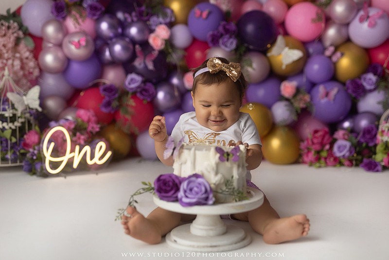 Colorful butterfly-themed first birthday cake smash featuring a happy baby girl enjoying birthday cake surrounded by vibrant purple, pink, and gold balloons, floral accents, butterflies, and a glowing "One" sign at Studio 120 Photography in Ramona, California.