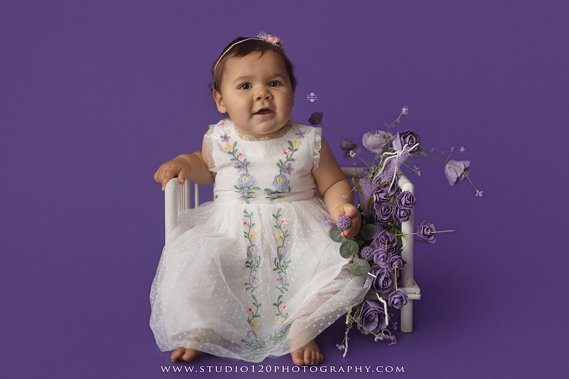 One-year milestone portraits of a smiling baby girl wearing a white embroidered dress and floral headband, seated in a modern white chair against a rich purple backdrop at Studio 120 Photography in Ramona, California.