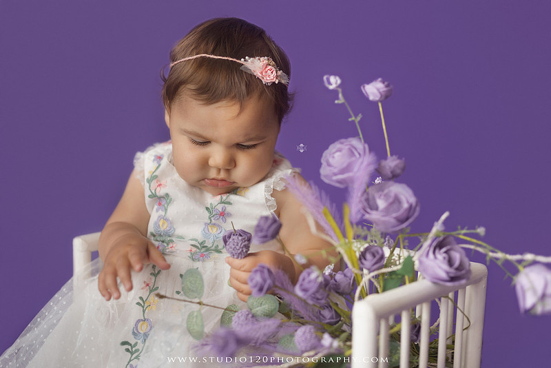 One-year milestone portraits of a smiling baby girl wearing a white embroidered dress and floral headband, seated in a modern white chair against a rich purple backdrop at Studio 120 Photography in Ramona, California.