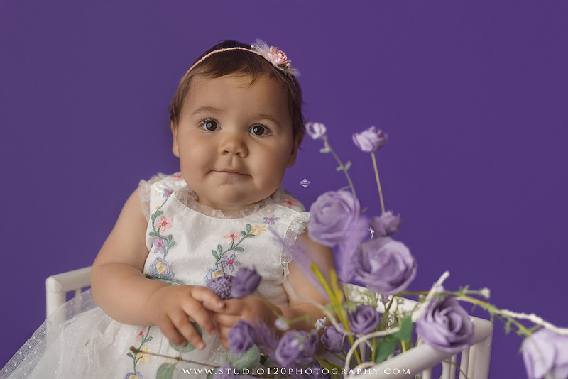 One-year milestone portraits of a smiling baby girl wearing a white embroidered dress and floral headband, seated in a modern white chair against a rich purple backdrop at Studio 120 Photography in Ramona, California.