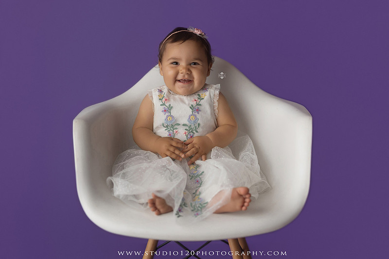 One-year milestone portraits of a smiling baby girl wearing a white embroidered dress and floral headband, seated in a modern white chair against a rich purple backdrop at Studio 120 Photography in Ramona, California.