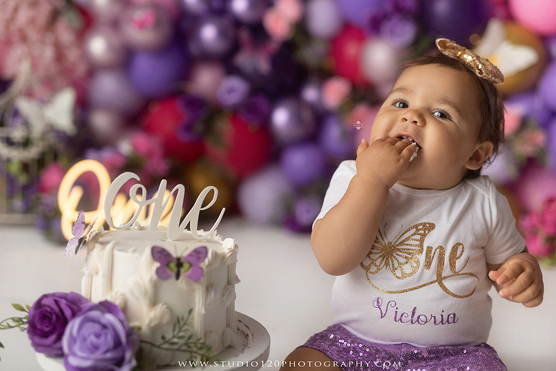 Colorful butterfly-themed first birthday cake smash featuring a happy baby girl enjoying birthday cake surrounded by vibrant purple, pink, and gold balloons, floral accents, butterflies, and a glowing "One" sign at Studio 120 Photography in Ramona, California.