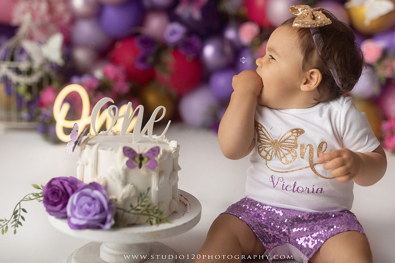 Colorful butterfly-themed first birthday cake smash featuring a happy baby girl enjoying birthday cake surrounded by vibrant purple, pink, and gold balloons, floral accents, butterflies, and a glowing "One" sign at Studio 120 Photography in Ramona, California.