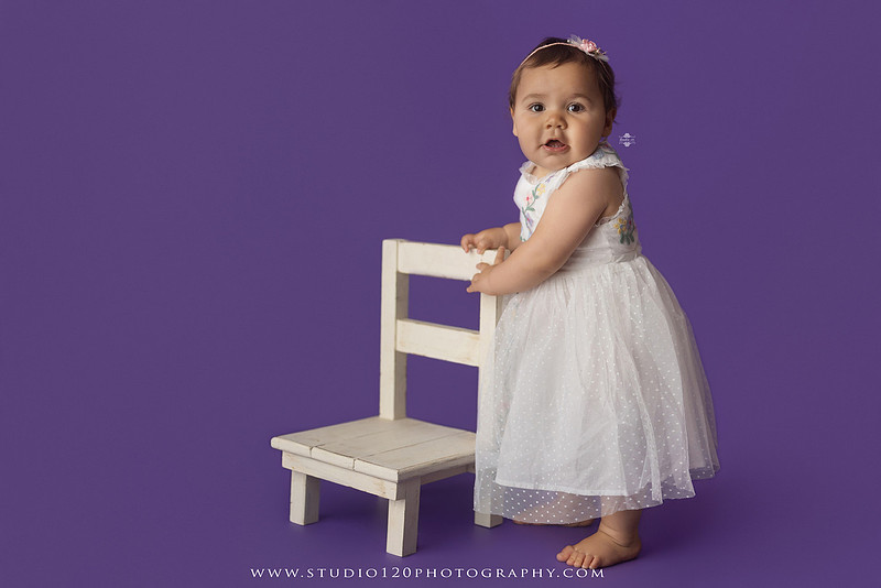 One-year milestone portraits of a smiling baby girl wearing a white embroidered dress and floral headband, seated in a modern white chair against a rich purple backdrop at Studio 120 Photography in Ramona, California.