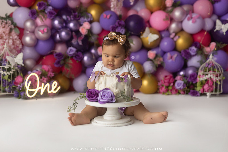 Colorful butterfly-themed first birthday cake smash featuring a happy baby girl enjoying birthday cake surrounded by vibrant purple, pink, and gold balloons, floral accents, butterflies, and a glowing "One" sign at Studio 120 Photography in Ramona, California.
