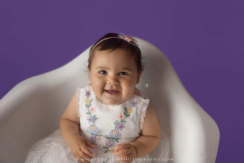 One-year milestone portraits of a smiling baby girl wearing a white embroidered dress and floral headband, seated in a modern white chair against a rich purple backdrop at Studio 120 Photography in Ramona, California.
