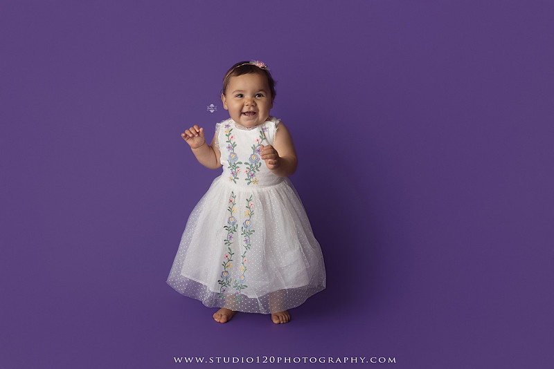 One-year milestone portraits of a smiling baby girl wearing a white embroidered dress and floral headband, seated in a modern white chair against a rich purple backdrop at Studio 120 Photography in Ramona, California.