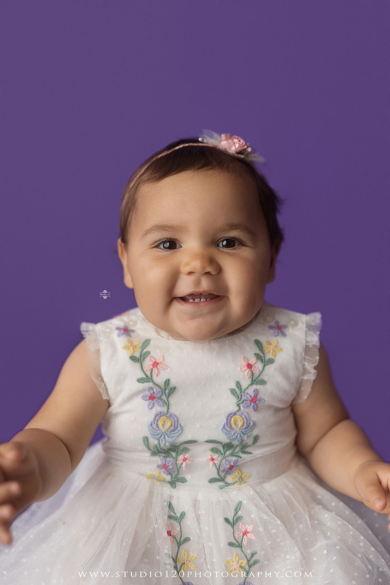 One-year milestone portraits of a smiling baby girl wearing a white embroidered dress and floral headband, seated in a modern white chair against a rich purple backdrop at Studio 120 Photography in Ramona, California.