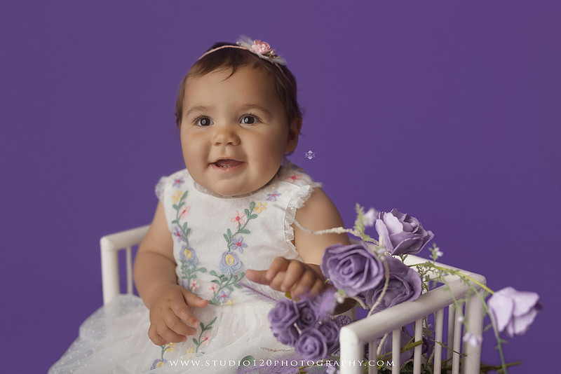 One-year milestone portraits of a smiling baby girl wearing a white embroidered dress and floral headband, seated in a modern white chair against a rich purple backdrop at Studio 120 Photography in Ramona, California.
