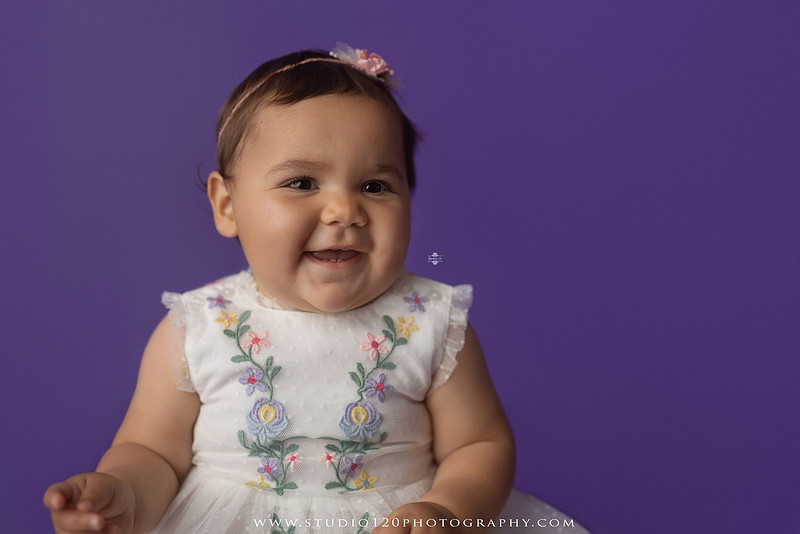 One-year milestone portraits of a smiling baby girl wearing a white embroidered dress and floral headband, seated in a modern white chair against a rich purple backdrop at Studio 120 Photography in Ramona, California.