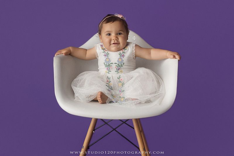 One-year milestone portraits of a smiling baby girl wearing a white embroidered dress and floral headband, seated in a modern white chair against a rich purple backdrop at Studio 120 Photography in Ramona, California.