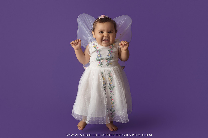 One-year milestone portraits of a smiling baby girl wearing a white embroidered dress and floral headband, seated in a modern white chair against a rich purple backdrop at Studio 120 Photography in Ramona, California.