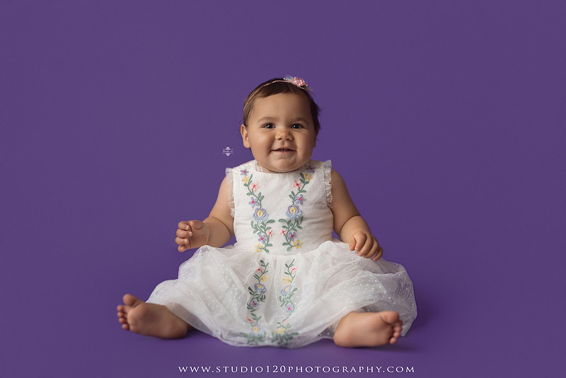 One-year milestone portraits of a smiling baby girl wearing a white embroidered dress and floral headband, seated in a modern white chair against a rich purple backdrop at Studio 120 Photography in Ramona, California.