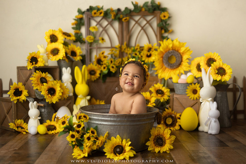 One-year-old baby girl enjoying a sunflower-themed milk bath in a vintage galvanized tub surrounded by bright yellow sunflowers, rustic spring decor, Easter accents, and cheerful floral details during a milestone photography session at Studio 120 Photography in Ramona, California.