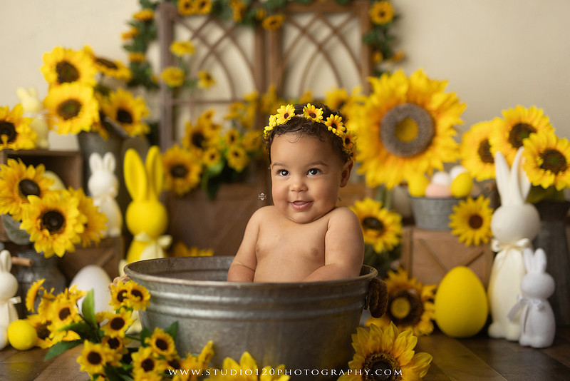One-year-old baby girl enjoying a sunflower-themed milk bath in a vintage galvanized tub surrounded by bright yellow sunflowers, rustic spring decor, Easter accents, and cheerful floral details during a milestone photography session at Studio 120 Photography in Ramona, California.