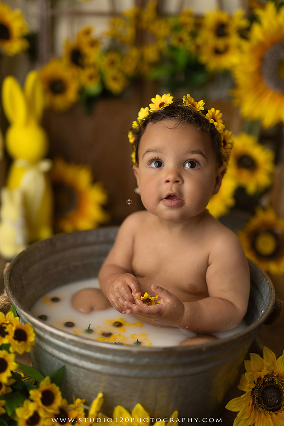 One-year-old baby girl enjoying a sunflower-themed milk bath in a vintage galvanized tub surrounded by bright yellow sunflowers, rustic spring decor, Easter accents, and cheerful floral details during a milestone photography session at Studio 120 Photography in Ramona, California.