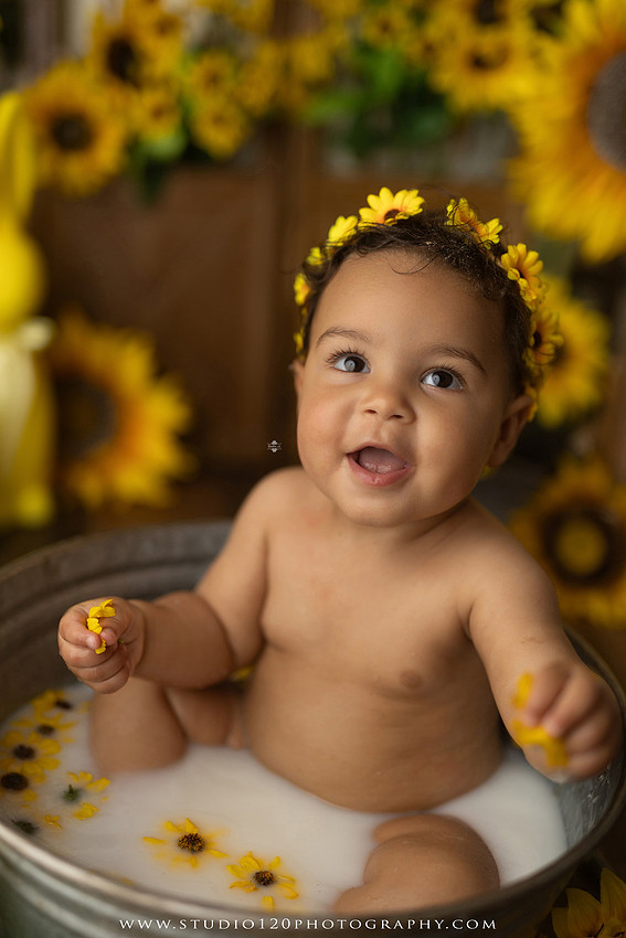 One-year-old baby girl enjoying a sunflower-themed milk bath in a vintage galvanized tub surrounded by bright yellow sunflowers, rustic spring decor, Easter accents, and cheerful floral details during a milestone photography session at Studio 120 Photography in Ramona, California.