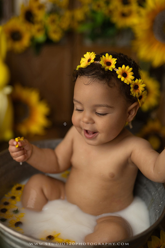 One-year-old baby girl enjoying a sunflower-themed milk bath in a vintage galvanized tub surrounded by bright yellow sunflowers, rustic spring decor, Easter accents, and cheerful floral details during a milestone photography session at Studio 120 Photography in Ramona, California.