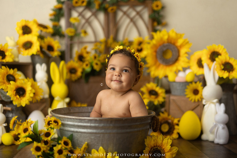 One-year-old baby girl enjoying a sunflower-themed milk bath in a vintage galvanized tub surrounded by bright yellow sunflowers, rustic spring decor, Easter accents, and cheerful floral details during a milestone photography session at Studio 120 Photography in Ramona, California.