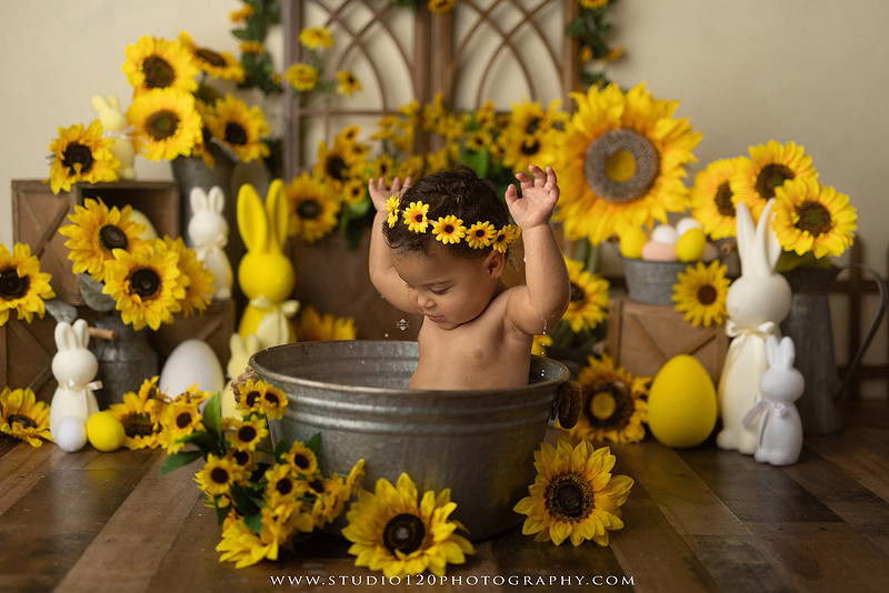 One-year-old baby girl enjoying a sunflower-themed milk bath in a vintage galvanized tub surrounded by bright yellow sunflowers, rustic spring decor, Easter accents, and cheerful floral details during a milestone photography session at Studio 120 Photography in Ramona, California.
