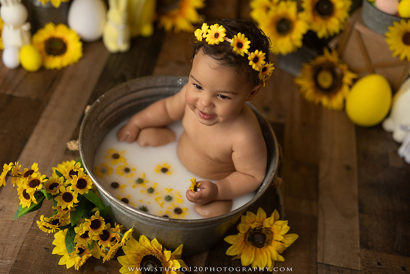 One-year-old baby girl enjoying a sunflower-themed milk bath in a vintage galvanized tub surrounded by bright yellow sunflowers, rustic spring decor, Easter accents, and cheerful floral details during a milestone photography session at Studio 120 Photography in Ramona, California.
