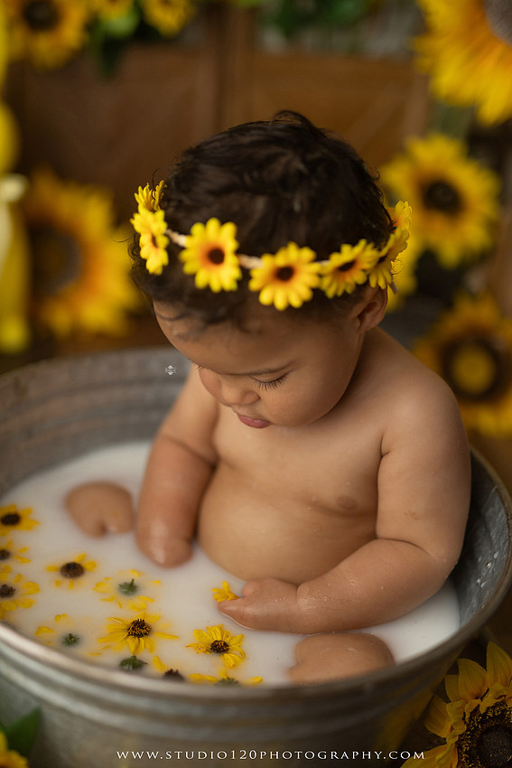 One-year-old baby girl enjoying a sunflower-themed milk bath in a vintage galvanized tub surrounded by bright yellow sunflowers, rustic spring decor, Easter accents, and cheerful floral details during a milestone photography session at Studio 120 Photography in Ramona, California.