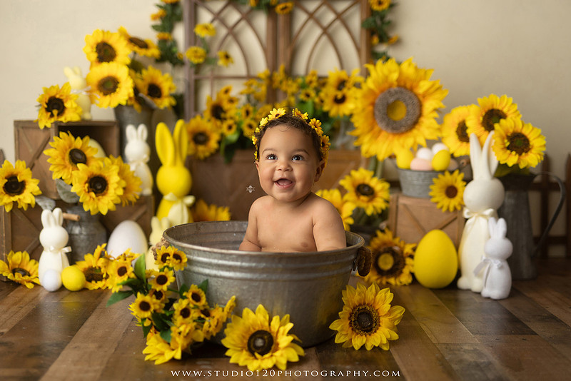 One-year-old baby girl enjoying a sunflower-themed milk bath in a vintage galvanized tub surrounded by bright yellow sunflowers, rustic spring decor, Easter accents, and cheerful floral details during a milestone photography session at Studio 120 Photography in Ramona, California.