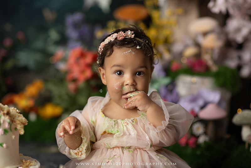 Baby girl enjoying a whimsical enchanted garden cake smash surrounded by oversized mushrooms, butterflies, blooming flowers, lush greenery, and a custom floral birthday cake while celebrating her first birthday at Studio 120 Photography in Ramona, California.