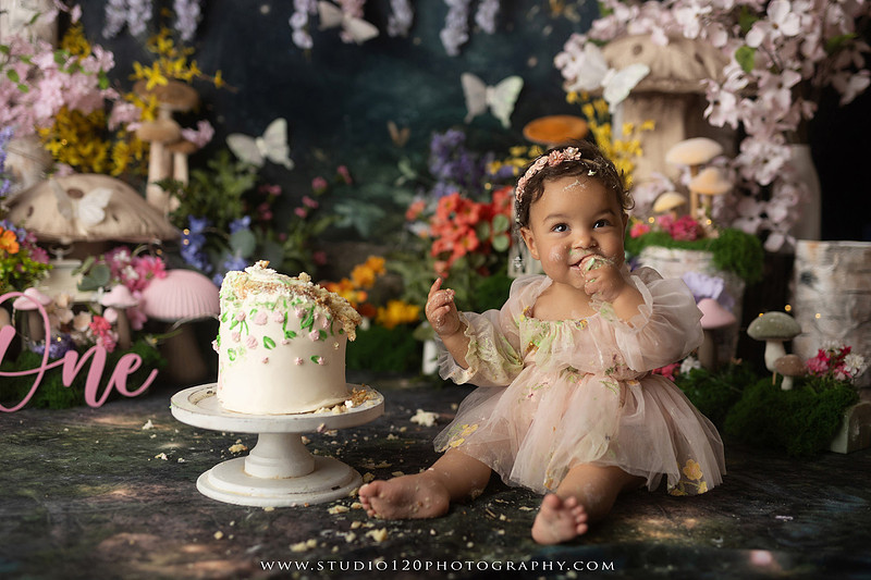 Baby girl enjoying a whimsical enchanted garden cake smash surrounded by oversized mushrooms, butterflies, blooming flowers, lush greenery, and a custom floral birthday cake while celebrating her first birthday at Studio 120 Photography in Ramona, California.