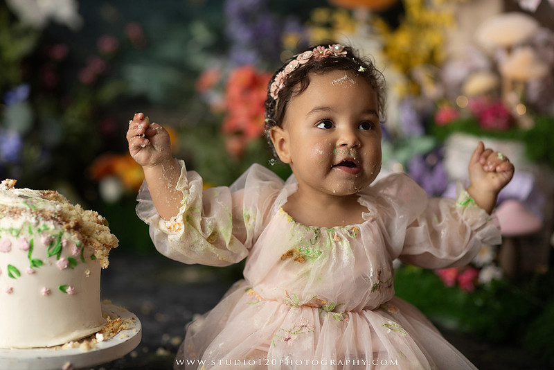 Baby girl enjoying a whimsical enchanted garden cake smash surrounded by oversized mushrooms, butterflies, blooming flowers, lush greenery, and a custom floral birthday cake while celebrating her first birthday at Studio 120 Photography in Ramona, California.
