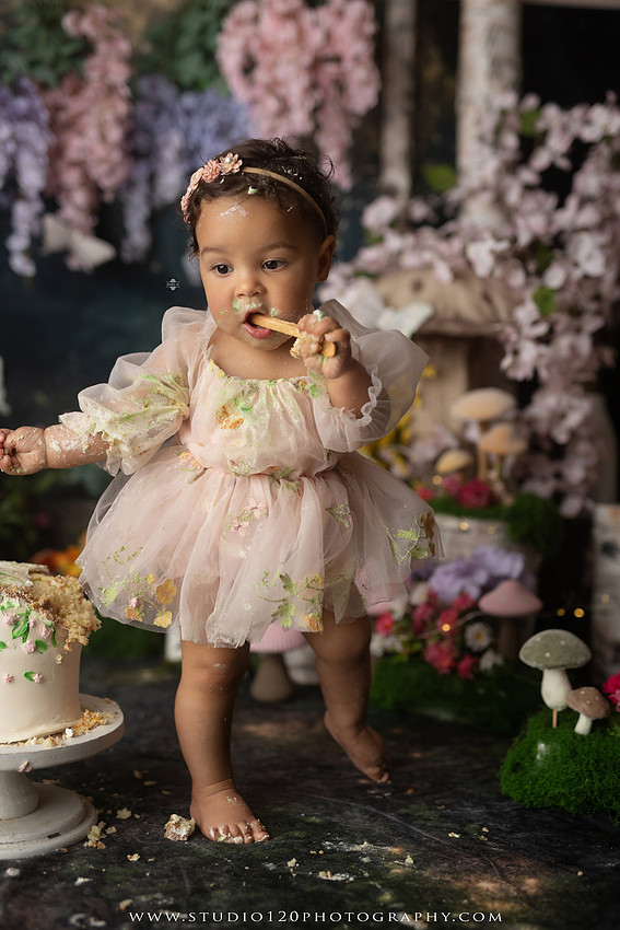Baby girl enjoying a whimsical enchanted garden cake smash surrounded by oversized mushrooms, butterflies, blooming flowers, lush greenery, and a custom floral birthday cake while celebrating her first birthday at Studio 120 Photography in Ramona, California.